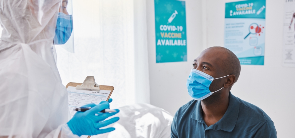 A man wearing a medical mask sites on a doctor's bed speaking to a healthcare professional in a full body protection suit with a blue medical mask and blue gloves.
